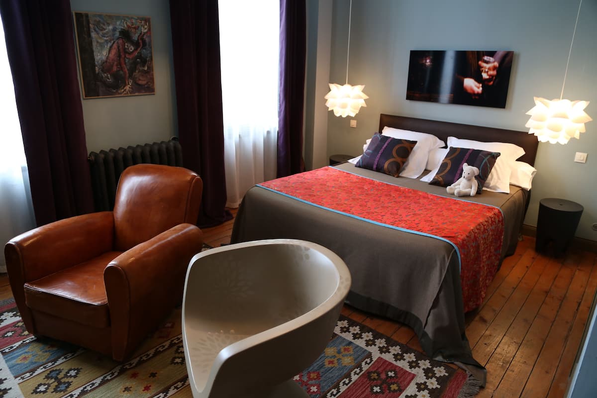 A hotel room with a bed, a brown leather armchair, a white chair, a patterned rug, and a radiator.