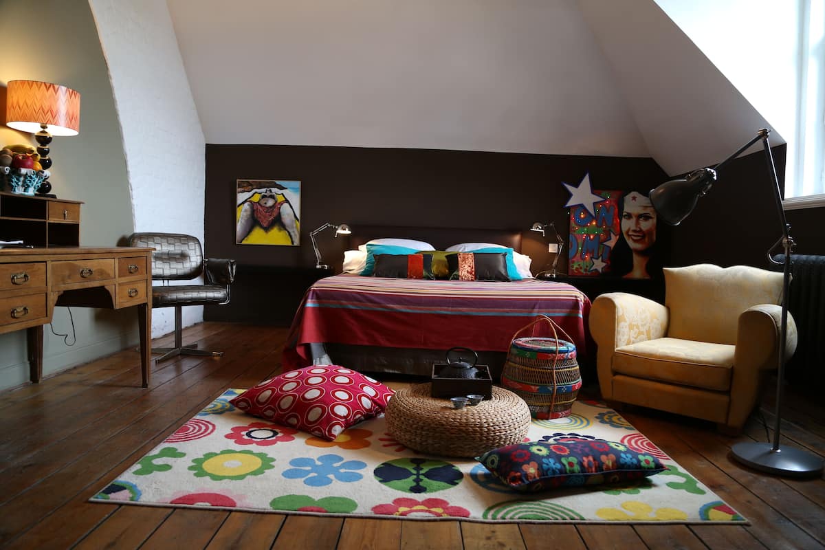 A bedroom with a sloped ceiling, a bed with colorful bedding, a chair, a desk, and a lamp.