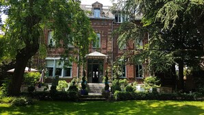 A historic brick building with a central entrance, surrounded by lush greenery and a well-maintained lawn.