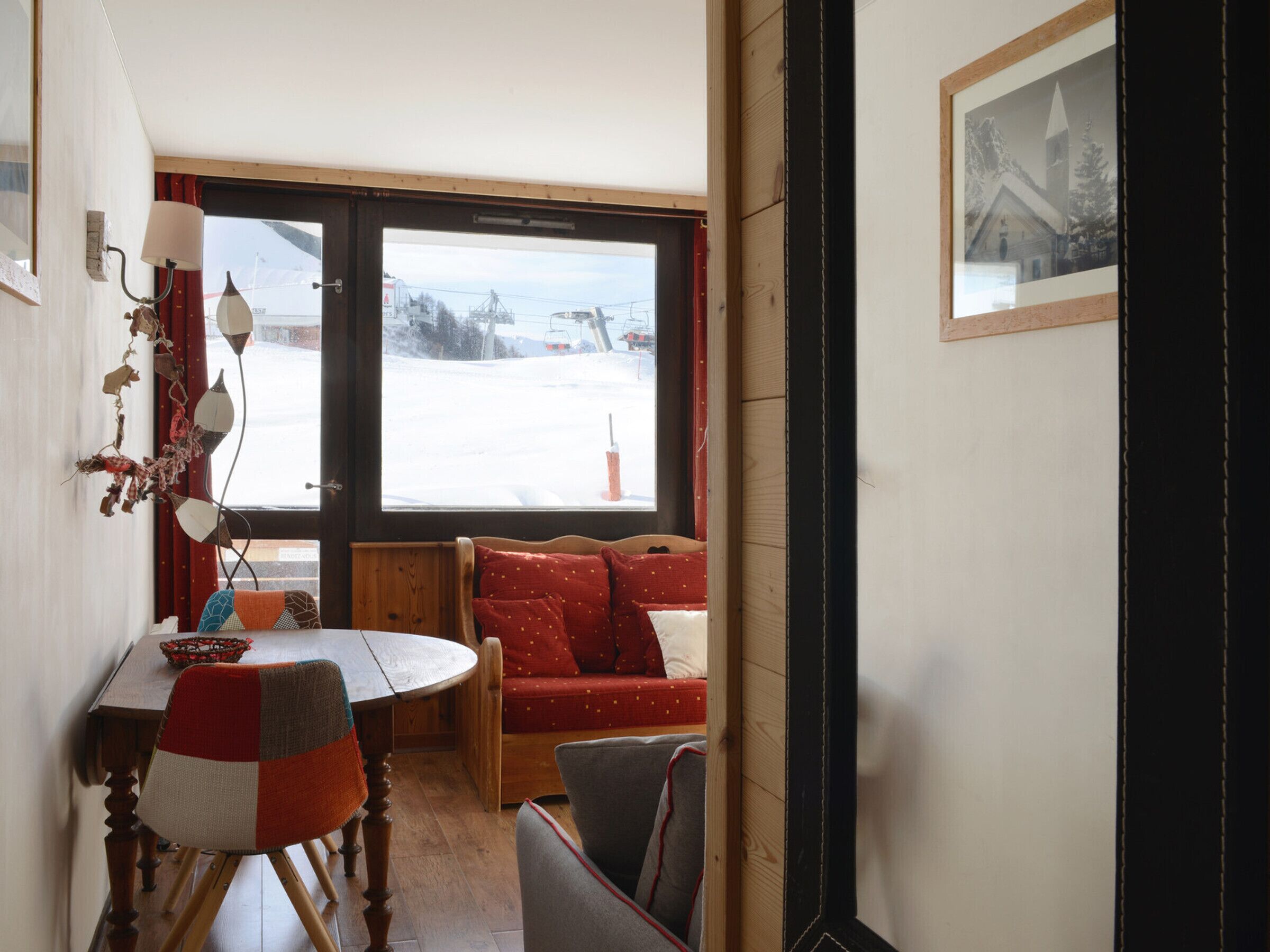 Ein gemütliches Zimmer mit einem Holzesstisch, einer Bank mit roten Kissen und einem großen Fenster mit Blick auf Schnee.