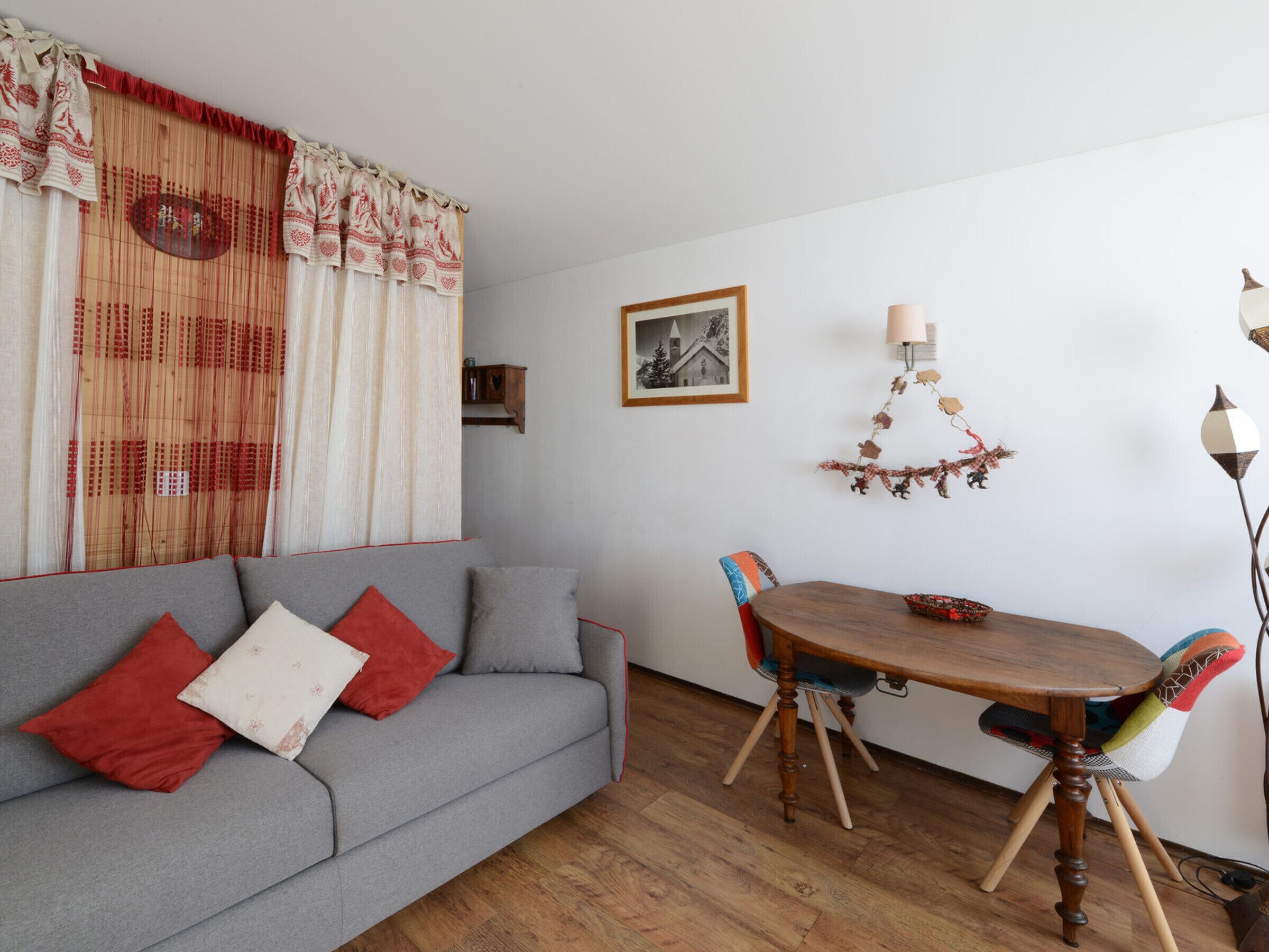 A cozy living area with a grey sofa, red and white cushions, a wooden dining table with chairs, and a wall-mounted lamp.