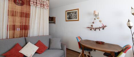 A cozy living area with a grey sofa, red and white cushions, a wooden dining table with chairs, and a wall-mounted lamp.