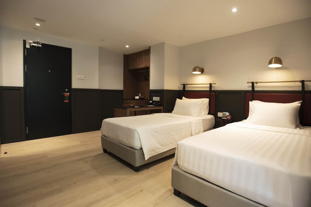 A hotel room with two beds, a wooden floor, a dark door, and a small table with a lamp.