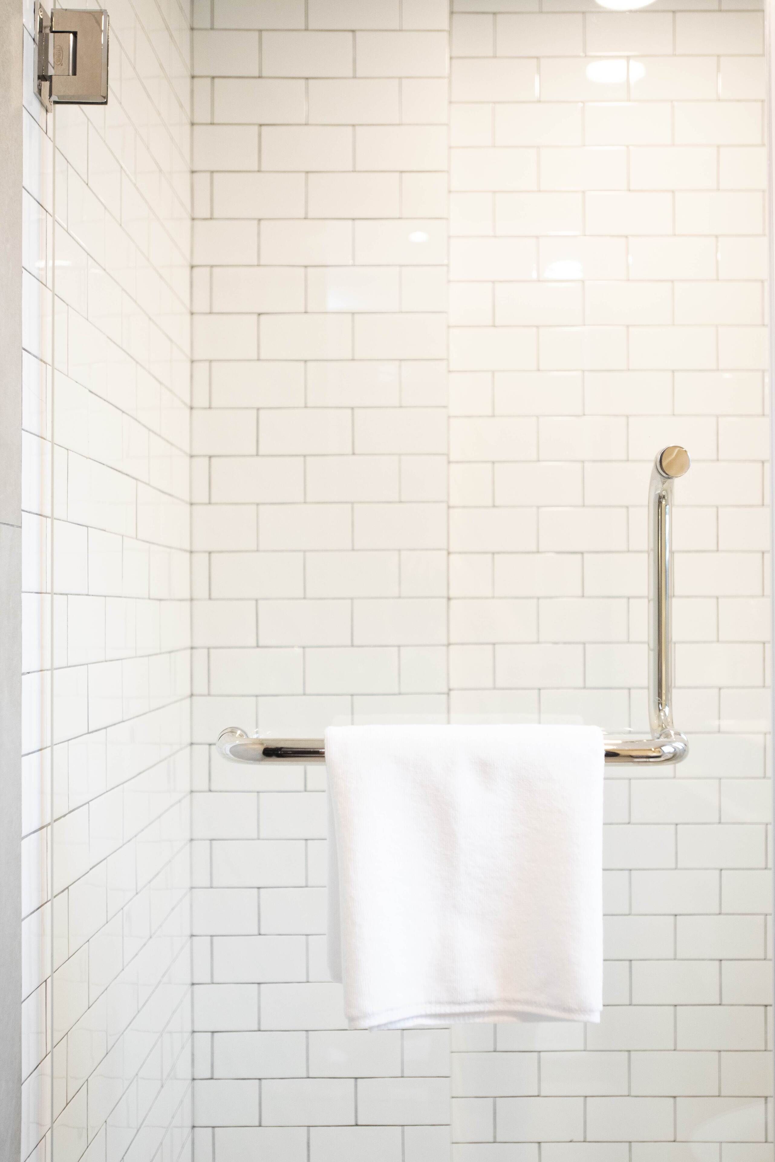 Un baño con paredes de azulejos blancos, una toalla blanca colgada en un perchero metálico y un pasamanos de cromo.