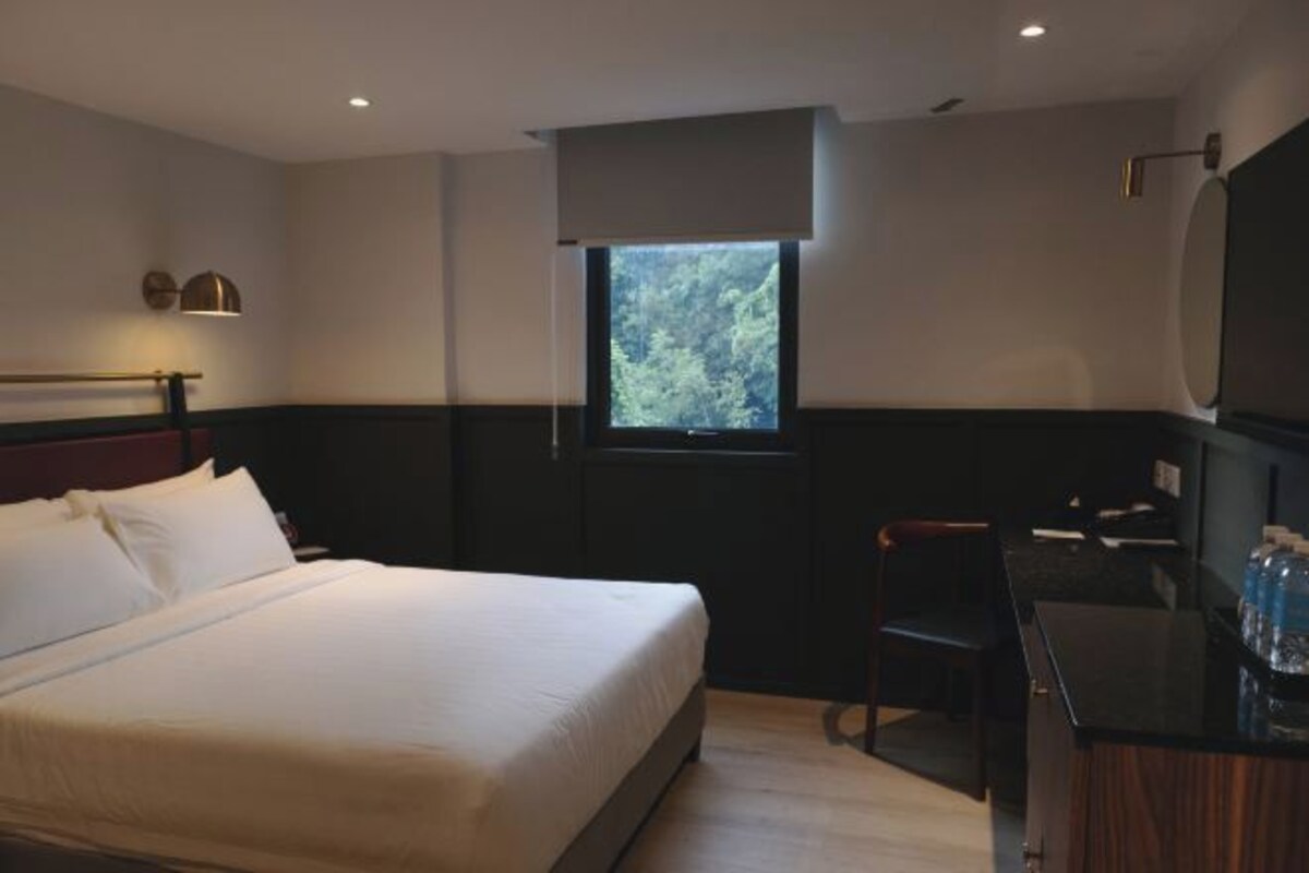 A hotel room with a bed, a desk, a chair, a window with a view of trees, and a mirror on the wall.