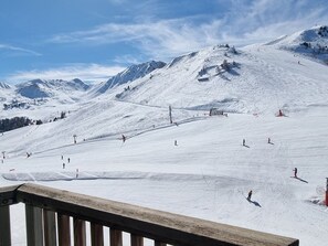 Pista de esqui coberta de neve com esquiadores, um deck de madeira e montanhas ao fundo.
