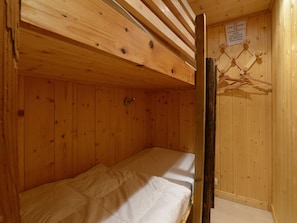 Ein kleiner, hölzerner Raum mit einem Etagenbett und einer Holzbalkendecke. An der Wand hängt ein Schild und ein Wäschegestell mit Kleidung.
