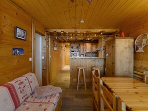 A wooden interior with a sofa, dining table, kitchen area, and a view of a mountain. - Cozy Apartment · Ski-in/Ski-out · 1 bedroom · Sleeps 6 · Mont Blanc Views (Plagne Aime 2000)