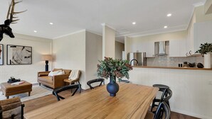 A modern living room with a wooden dining table, a sofa, and a coffee table. There is a kitchen area with white cabinets and stainless steel appliances. - Snowgums 17 - Dinner Plain (Dinner Plain)