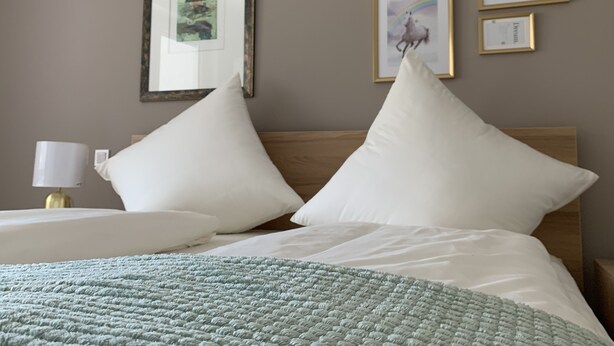 A neatly made bed with white pillows and a green blanket, a bedside lamp, and framed artwork on the wall. - Moderne Ferienwohnung mitten im Grünen in der Südeifel (Irrel)