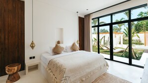A modern bedroom with a large bed, a wooden accent wall, and a view of a garden. - Villa Lamer (Canggu)