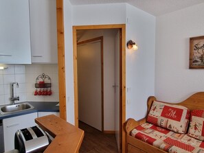 A compact living space with a kitchenette, a wooden bench, a sofa with a patterned cushion, and a door leading to another room.