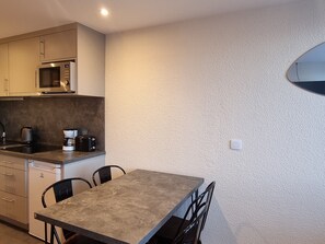 Uma cozinha compacta com micro-ondas, cafeteira e torradeira na bancada. Há uma mesa de jantar com cadeiras.