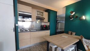 A compact kitchen with a wooden table, white stools, and a bunk bed in the background.