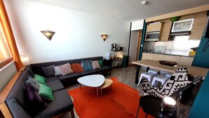 A modern living room with a sofa, coffee table, and patterned chairs. A kitchen area with appliances and a dining table is visible in the background.