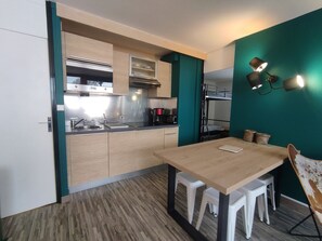 A compact kitchen with a wooden table, white stools, and a bunk bed in the background.