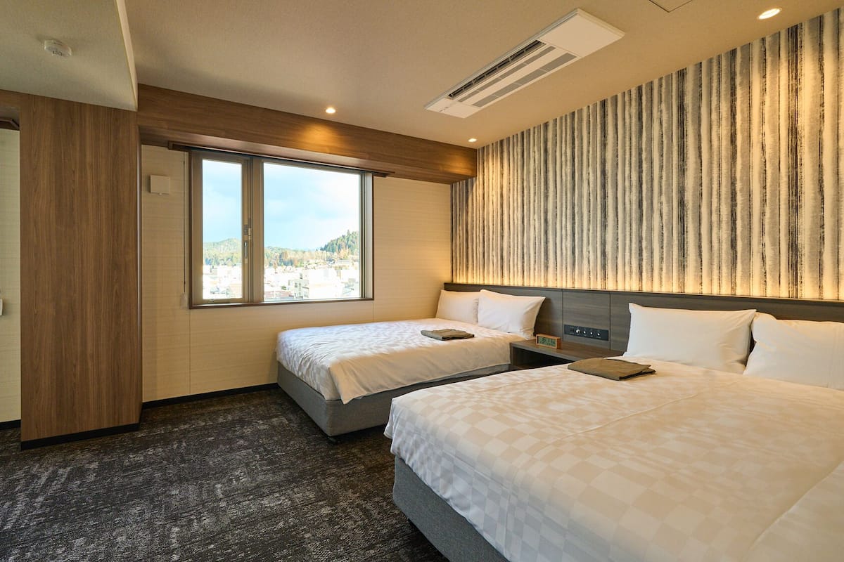 Habitación de hotel con dos camas, un ventanal grande, aire acondicionado de pared y vista a un paisaje urbano.