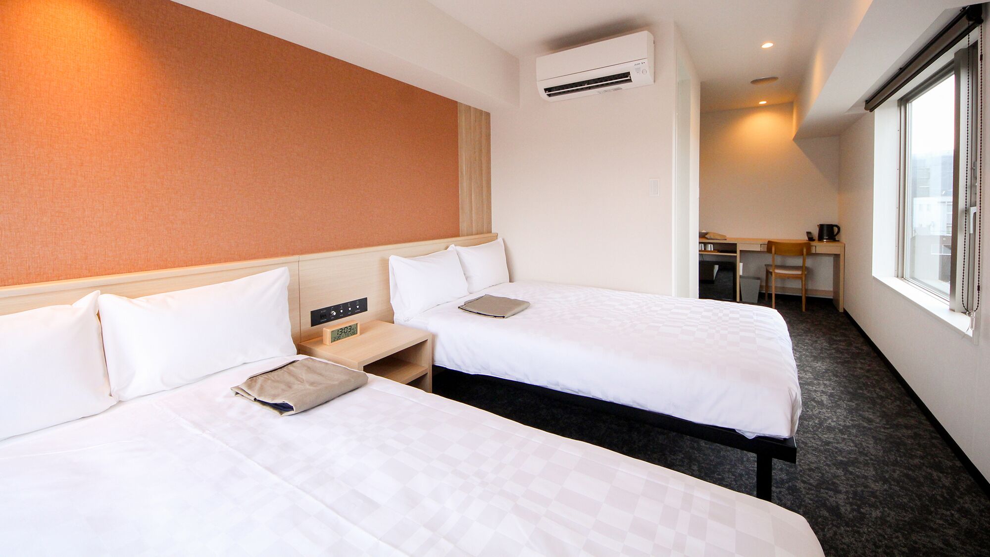 A hotel room with two beds, a desk, a chair, an air conditioning unit, and a window.