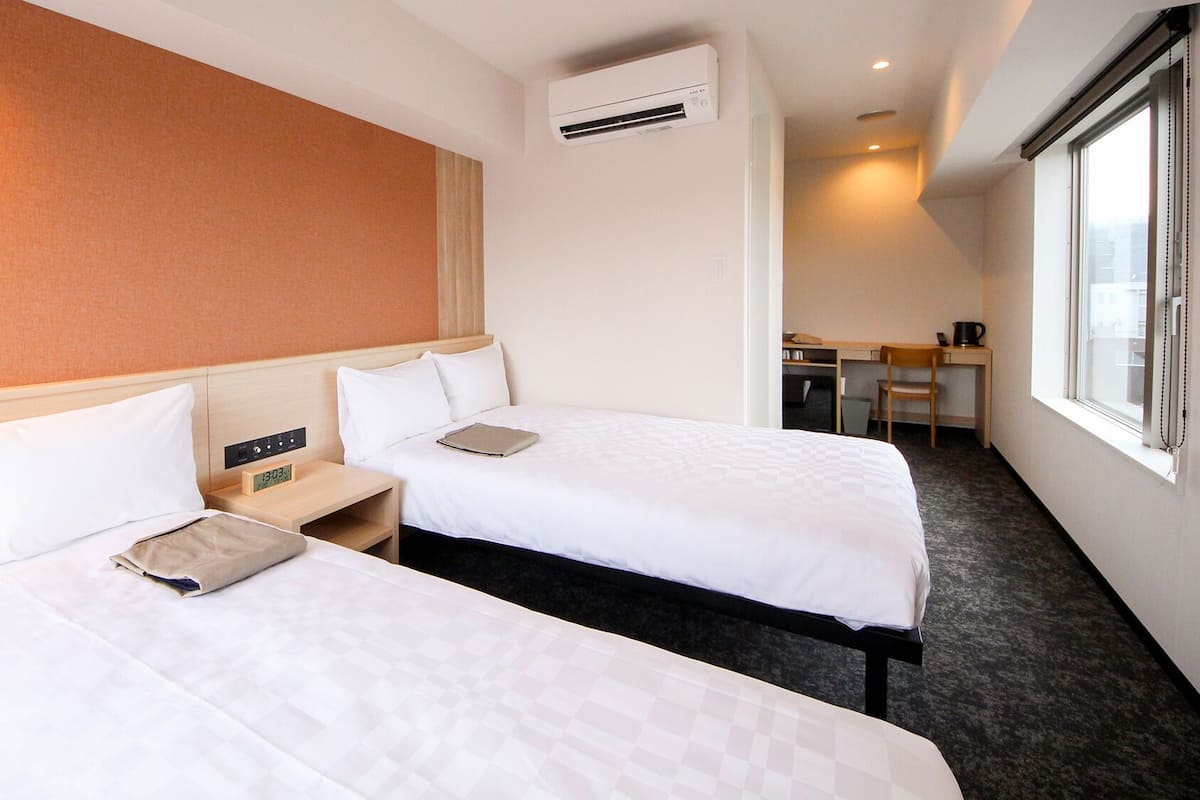 Habitación de hotel con dos camas, un escritorio, una silla, un equipo de aire acondicionado y una ventana.
