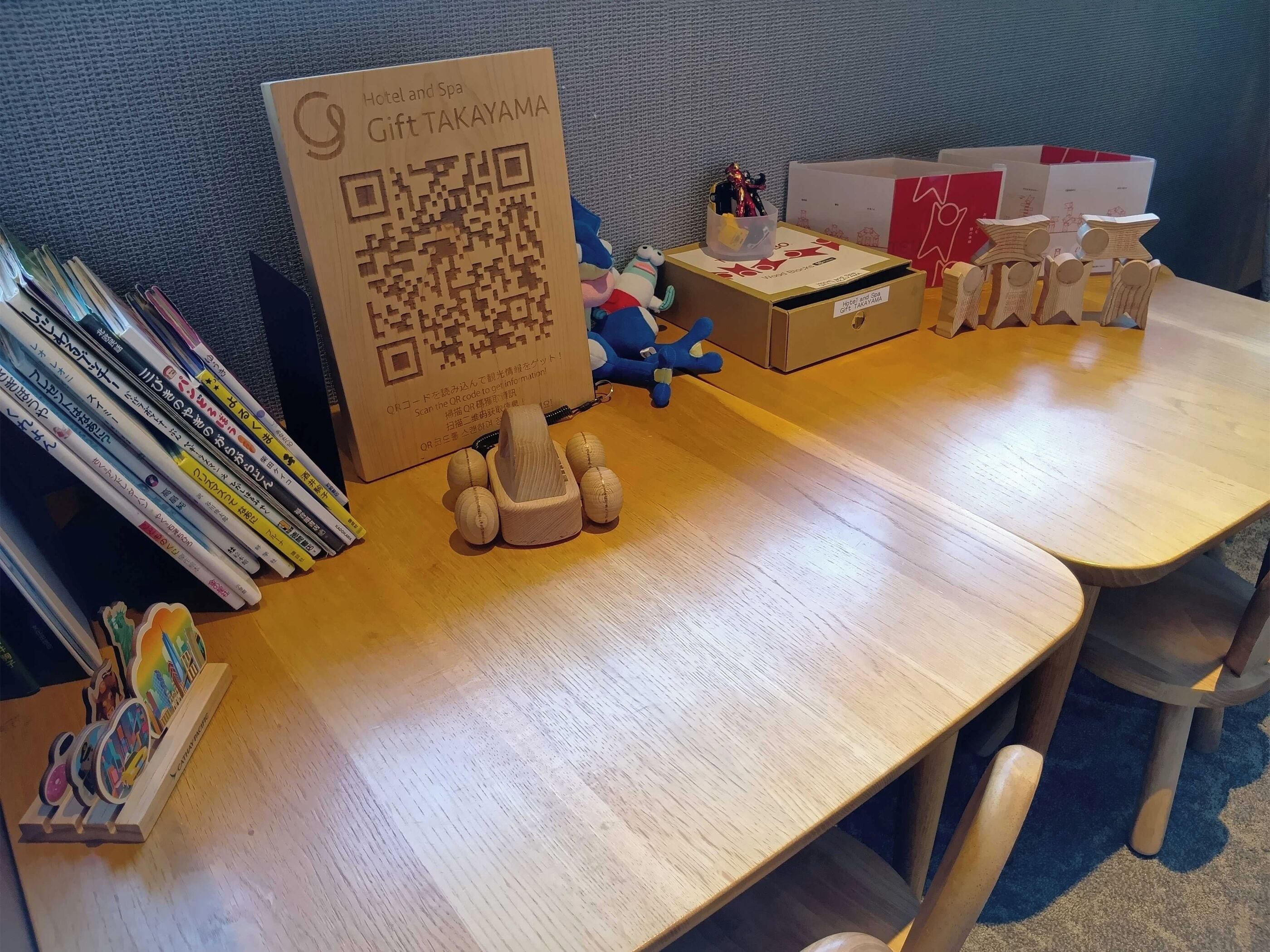 Puisesta pöydästä löytyy QR-koodilla varustettu kyltti, pino kirjoja, sininen pehmolelu ja puisia koristehahmoja.