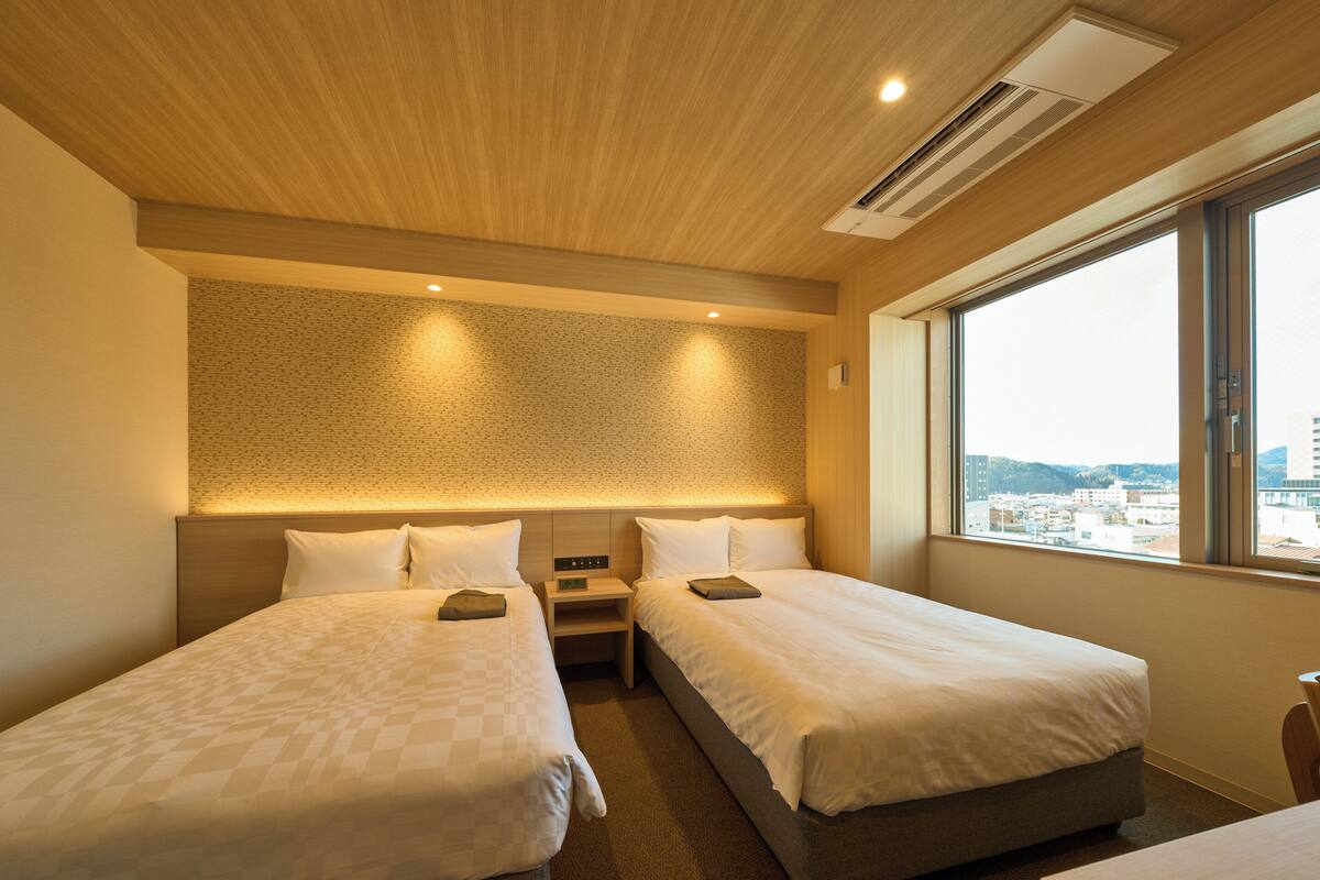 Habitación de hotel con dos camas, un ventanal grande y vistas a la ciudad.