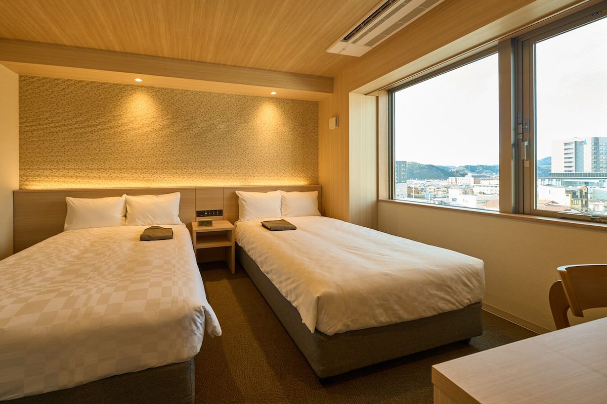 Habitación de hotel con dos camas, una mesita, una silla, una mesita de noche y vistas a la ciudad desde la ventana.