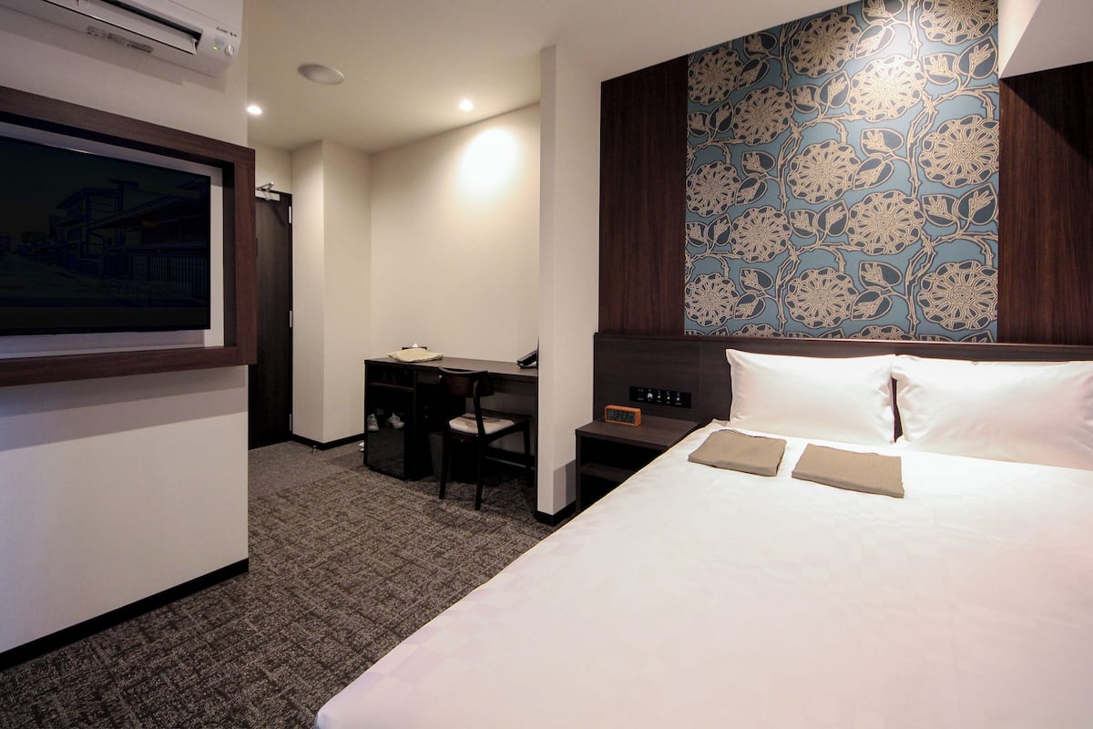 Habitación de hotel con una cama, un escritorio y una silla. Hay un reloj en la mesita de noche y un equipo de aire acondicionado en la pared.