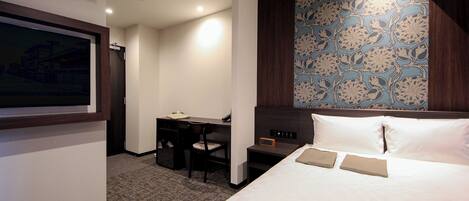 A hotel room with a bed, a desk, and a chair. There is a clock on the nightstand and an air conditioning unit on the wall.