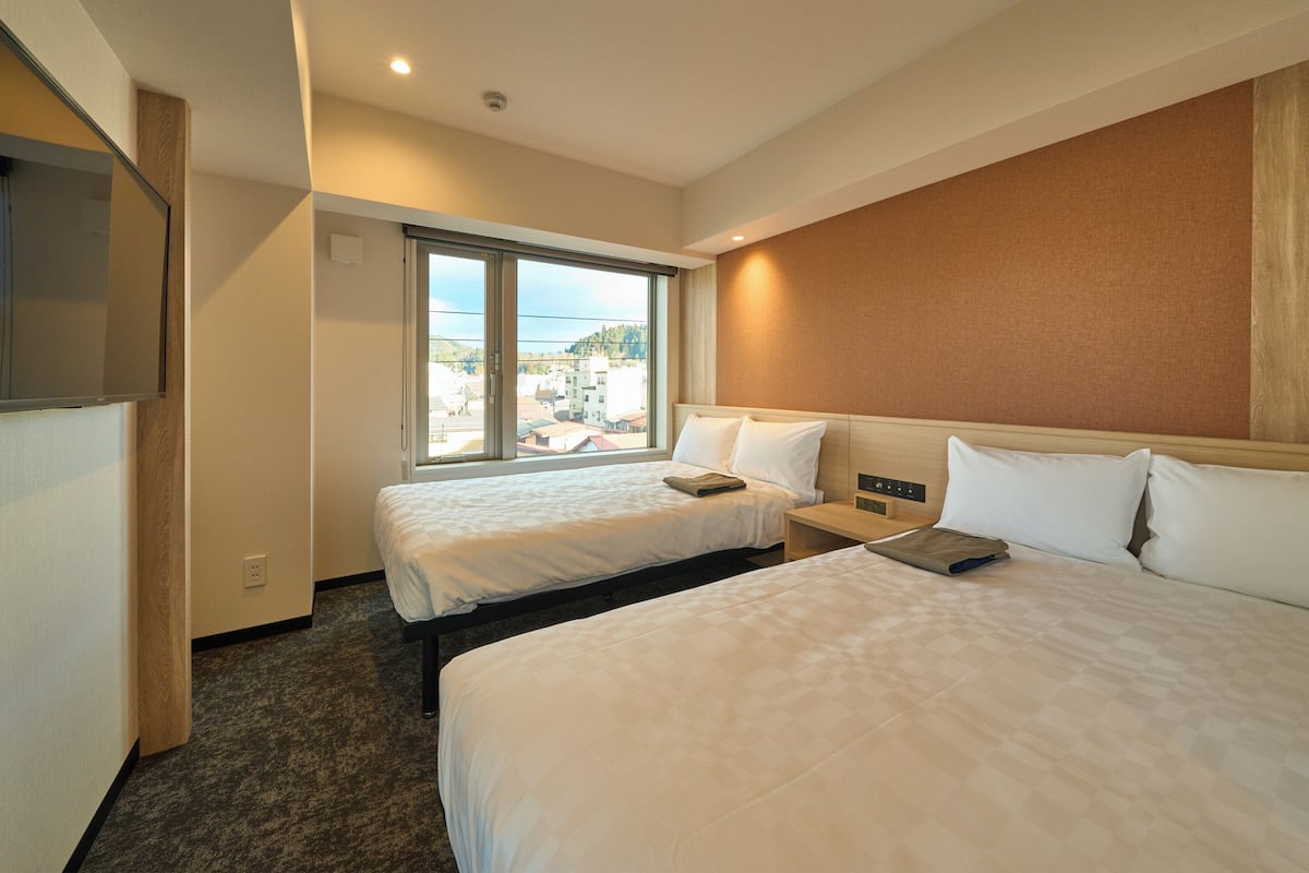 Habitación de hotel con dos camas, un ventanal con vistas a edificios, un televisor de pantalla plana montado en la pared y un cabecero de madera.