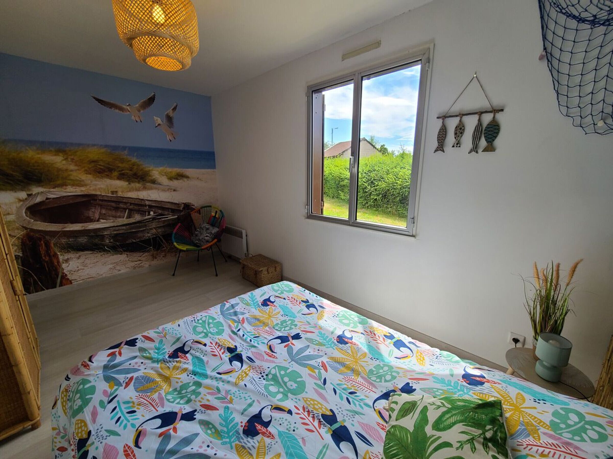 Un dormitorio con un mural de playa, un ventanal grande, una colcha colorida y un sillón hamaca.