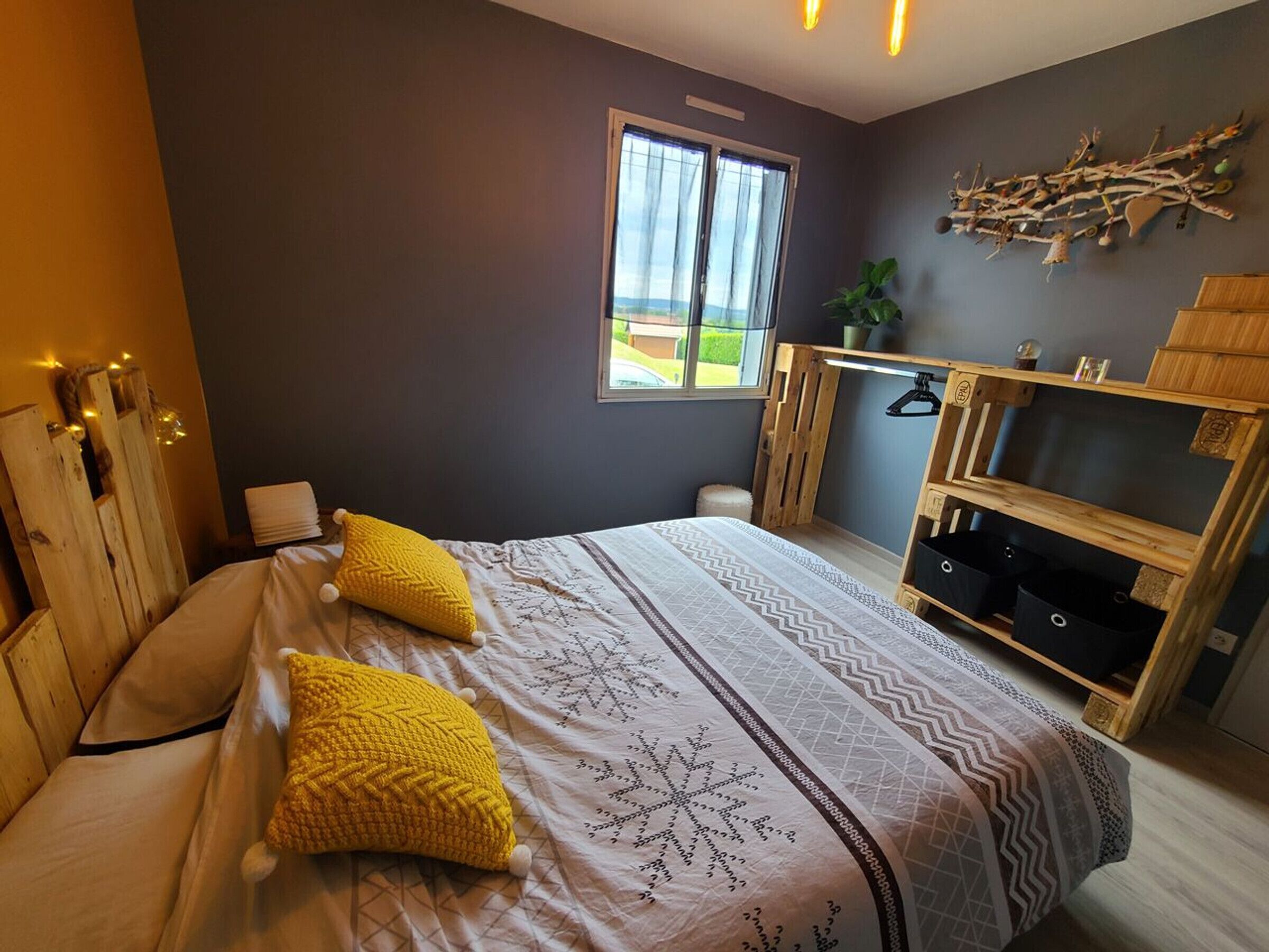 Un dormitorio con cama, dos almohadas, un armario de madera, una ventana y una planta.