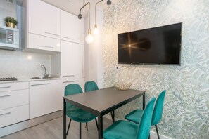 A modern kitchen with white cabinets, a built-in microwave, and a sink. There is a dining table with teal chairs and a flat-screen TV mounted on the wall.