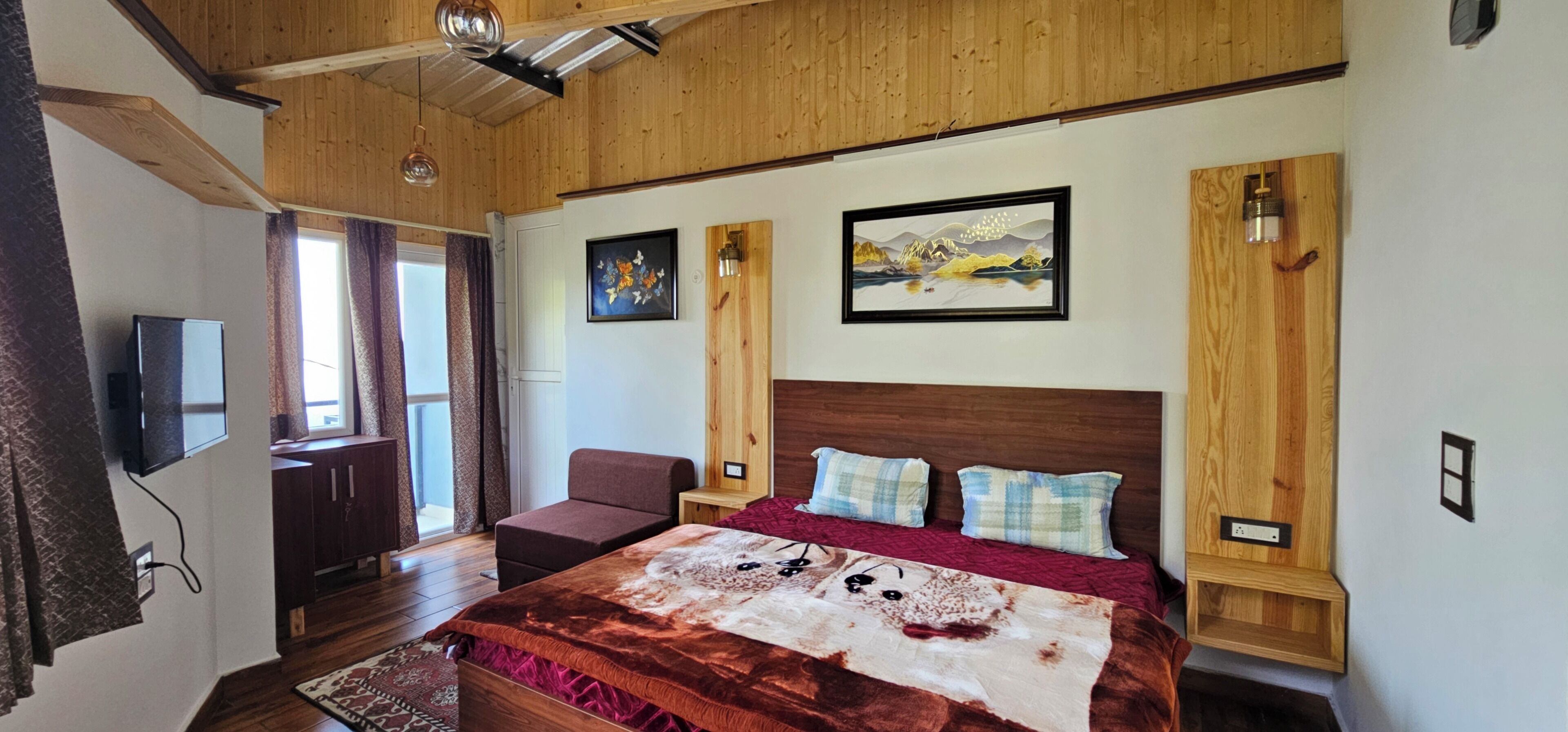 Une chambre avec un plafond en bois, un lit recouvert d’une couverture rouge et marron, deux oreillers, un téléviseur fixé au mur et deux lampes murales.