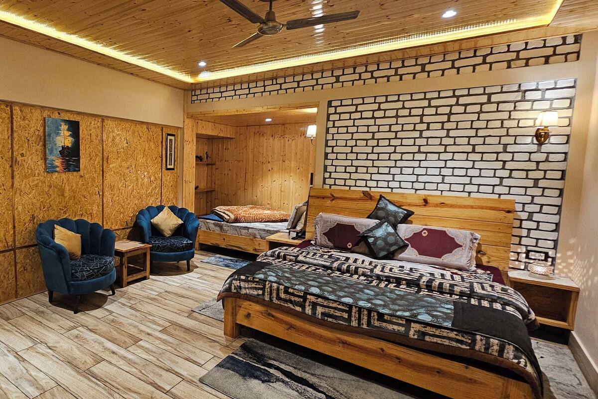 Ein Zimmer mit Holzboden, einem Bett mit gemusterten Textilien, zwei blauen Sesseln, einem kleinen Tisch und einer Wand mit Backsteinmuster.