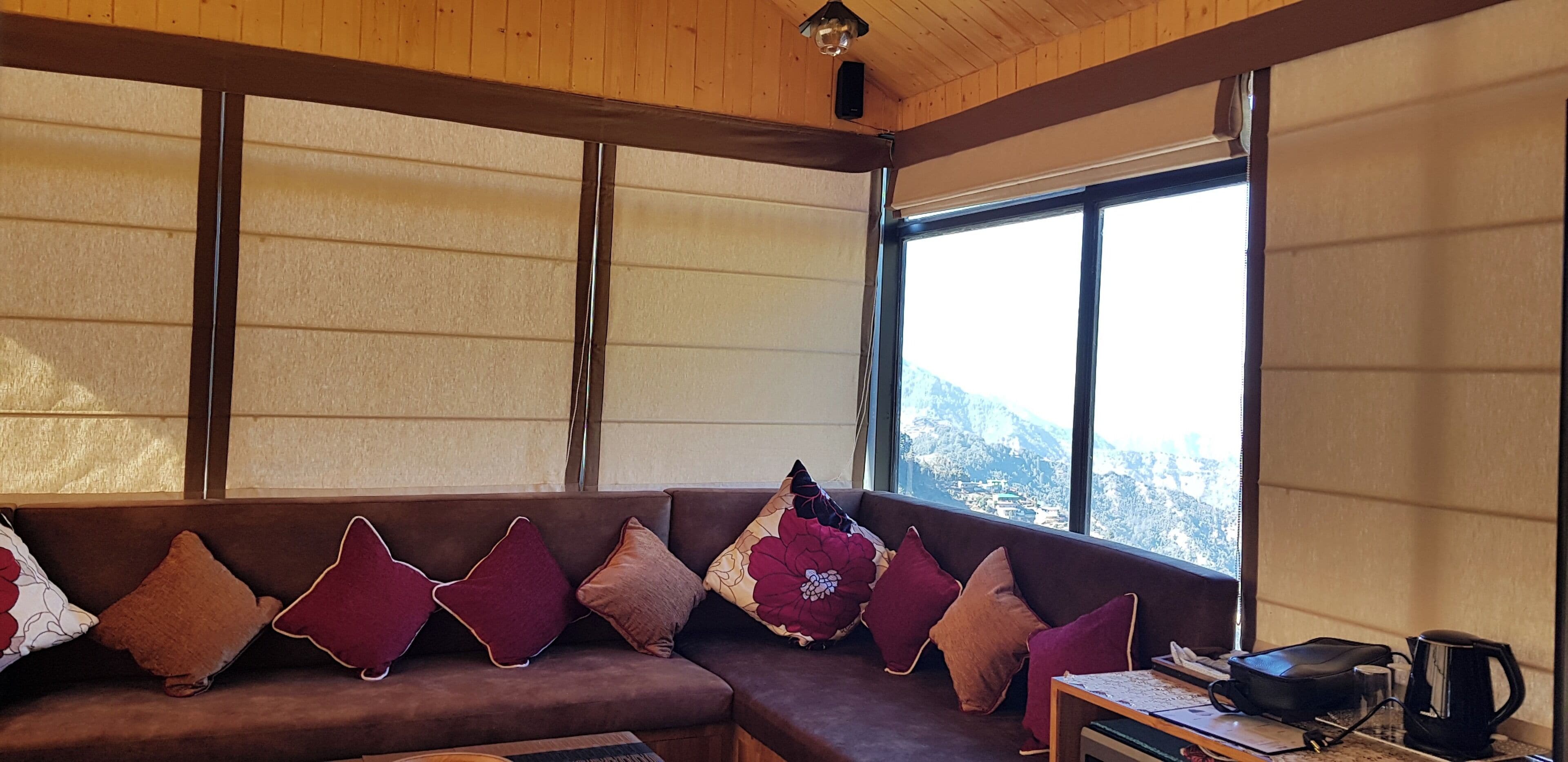 Um quarto acolhedor com um sofá marrom decorado com vários almofados, teto de madeira e uma grande janela com vista para as montanhas.