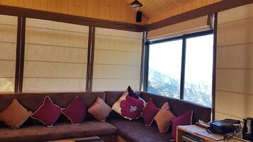 Um quarto acolhedor com um sofá marrom decorado com vários almofados, teto de madeira e uma grande janela com vista para as montanhas.