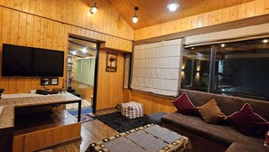 Una habitación acogedora con paredes de madera, un televisor de pantalla plana, un sofá con cojines, una mesita y una ventana con persianas.
