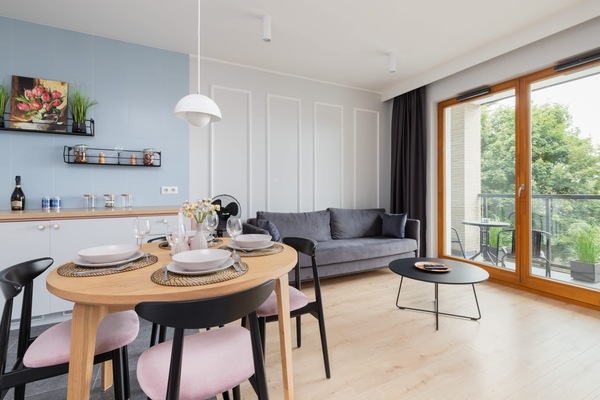 A modern living room with a dining table, chairs, a gray sofa, and large windows that let in natural light. The space features stylish shelves and decor elements.