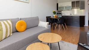 A modern living room with a grey sofa, two round wooden side tables, and a dining area with a black table and chairs. A kitchen with white cabinets and a microwave is visible in the background.
