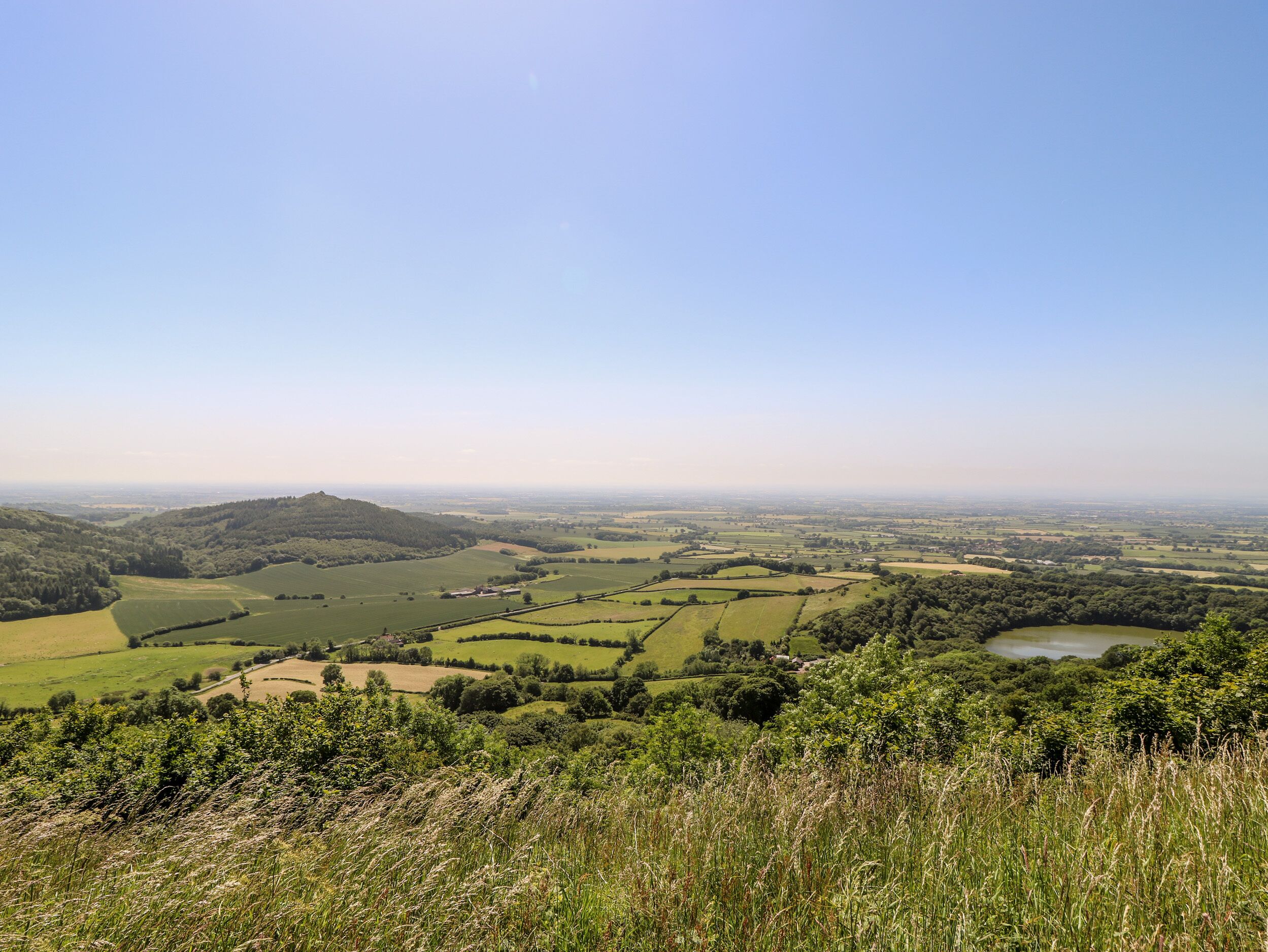 Ein weitreichender Panoramablick auf eine ländliche Landschaft mit sanften Hügeln, Feldern und einem kleinen Gewässer.