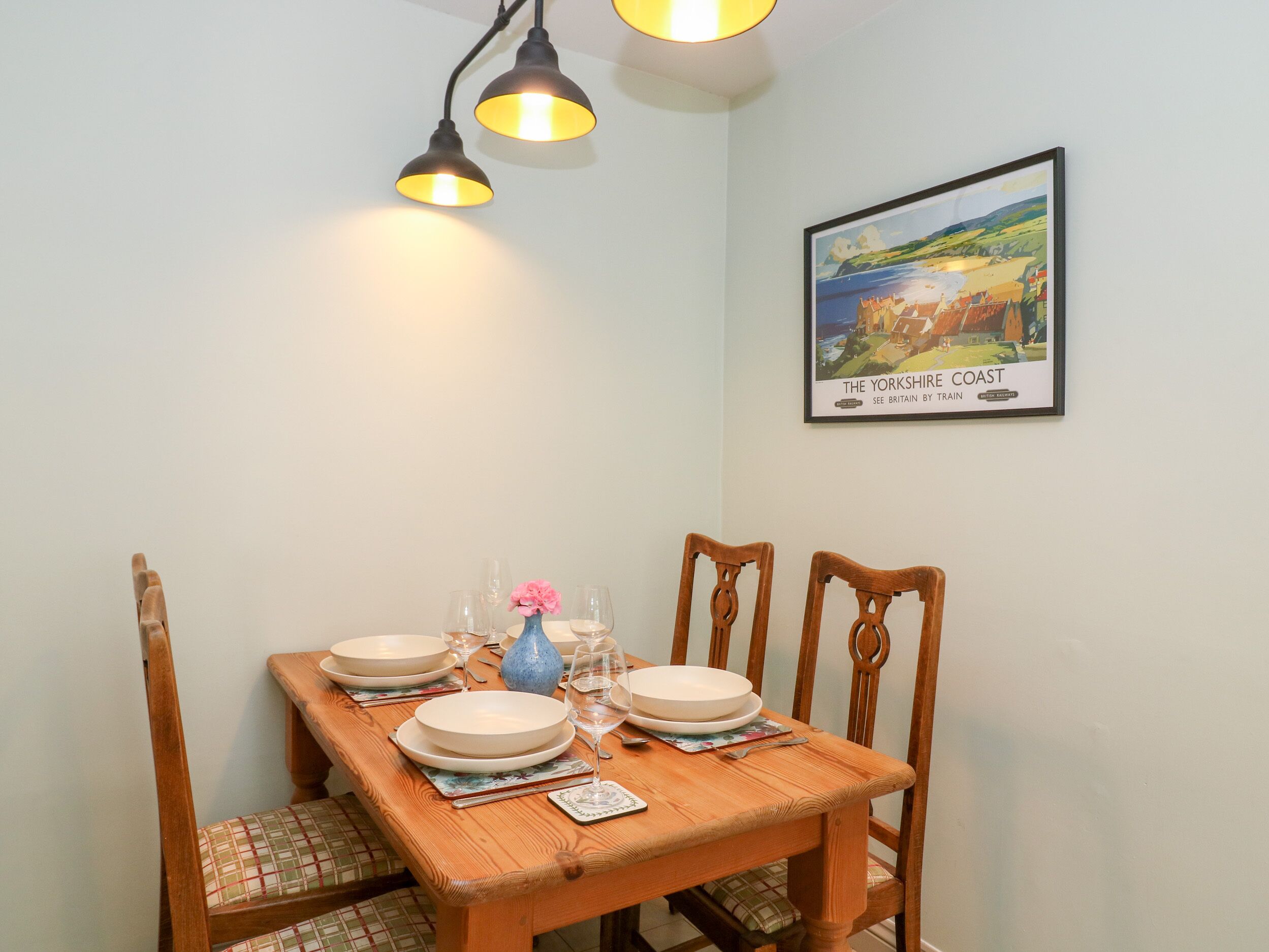 Una mesa de comedor dispuesta con platos, vasos y un florero, rodeada de sillas.