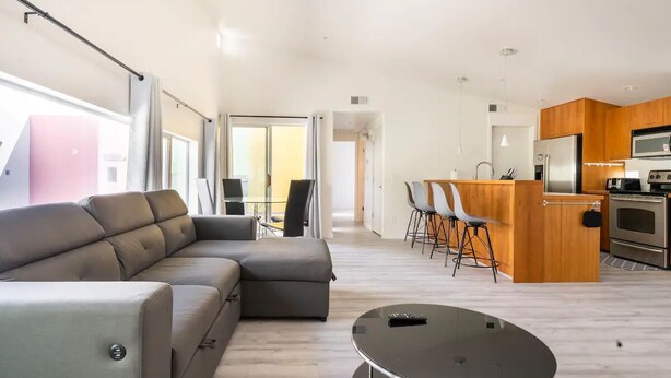 Een moderne woonkamer met een grijze modulaire bank, een ronde salontafel en een eethoek met een houten bar en stoelen.