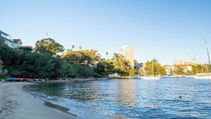 A beach with a sandy shore, a calm sea, and buildings in the background. - Getawayz NSW - Bay Court III -Spacious Waterside Studio next to Hayes Beach, Cafes & Ferry! (Neutral Bay)