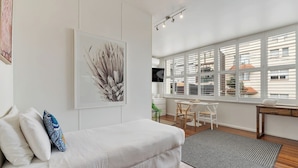 A modern living room with a white sofa, a dining area with a table and chairs, and large windows with blinds. - Getawayz NSW - Bay Court III -Spacious Waterside Studio next to Hayes Beach, Cafes & Ferry! (Neutral Bay)