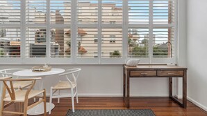 A room with a large window, a round table, two chairs, a wooden desk, and a rug. - Getawayz NSW - Bay Court III -Spacious Waterside Studio next to Hayes Beach, Cafes & Ferry! (Neutral Bay)