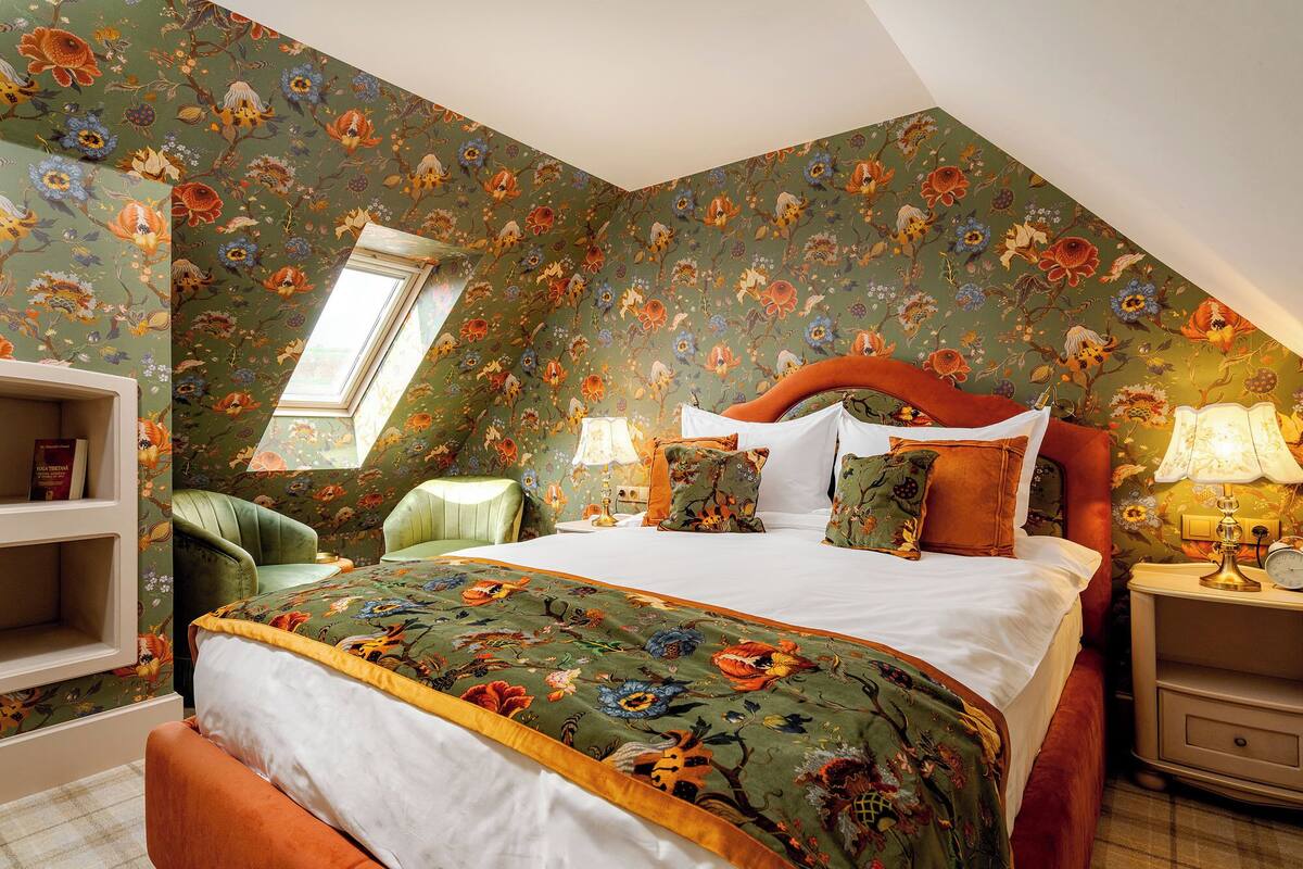 A bedroom with a large bed, a nightstand, a chair, a bookshelf, and a window.