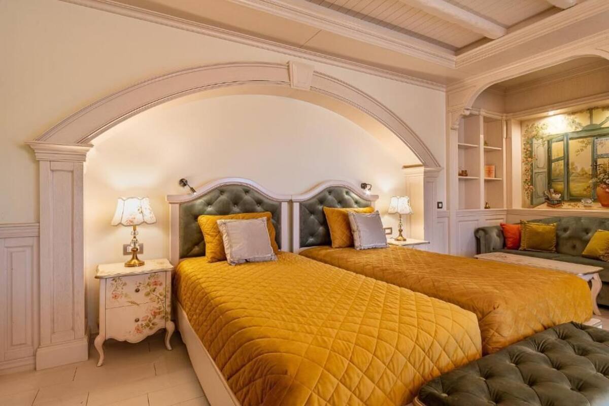 A bedroom with two beds, each with a tufted headboard and yellow bedding. There are nightstands with lamps and a green upholstered bench.