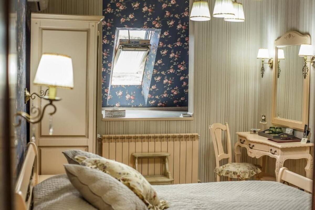 A bedroom with a bed, a dresser, a chair, a window with floral curtains, and wall-mounted lamps.