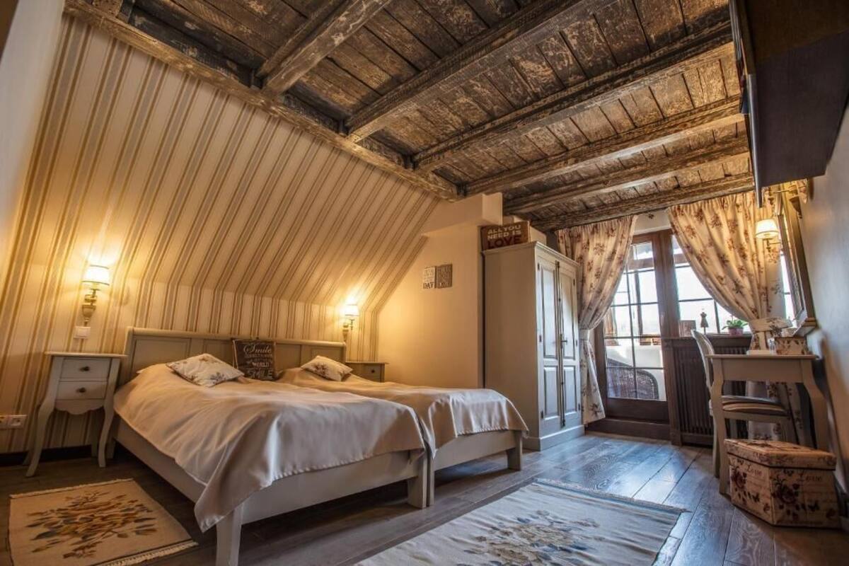 A bedroom with two beds, a wooden ceiling, a window with curtains, and a small table with a lamp.