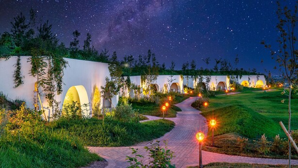 A pathway leading to a row of white buildings with arches, surrounded by greenery and illuminated by star-shaped lights.